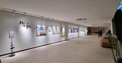 1. [표 좌측] 스타필드 수원 작은미술관 현장 사진, 에스컬레이터 좌측으로 길다란 통로가 있음. 좌측 흰색 벽면에 작품이 다수 걸려있음. 우측에는 등받이 없는 소파가 2개 놓아져 있음.